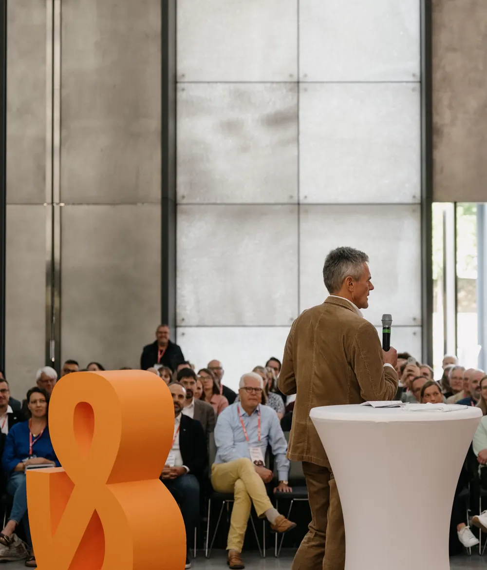 Ein Redner steht vor dem Publikum der TALER & TALAR Konferenz. Neben ihm steht ein großes orangefarbenes Und-Zeichen.